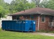 Blue dumpster in a residential driveway beside a brick house with a brown roof and green trees in the yard.