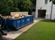 Blue dumpster labeled 'PEEPS Containers' filled with wood and cardboard debris on a driveway beside a white brick house with an open garage door.
