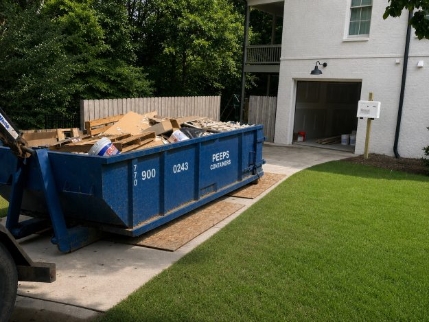 residential-renovation-dumpster-rental-atlanta-peeps-containers Blue dumpster labeled 'PEEPS Containers' filled with wood and cardboard debris on a driveway beside a white brick house with an open garage door.