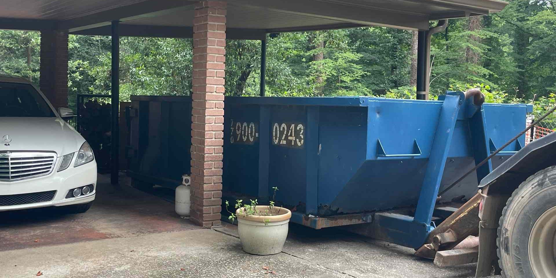 basement-cleanout-dumpster-rental-brookhaven-ga White Mercedes under a carport next to a large blue dumpster, with brick columns and greenery in the background.