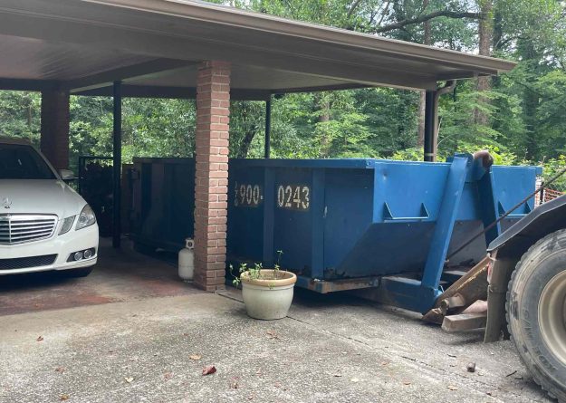 basement-cleanout-dumpster-rental-brookhaven-ga White Mercedes under a carport next to a large blue dumpster, with brick columns and greenery in the background.