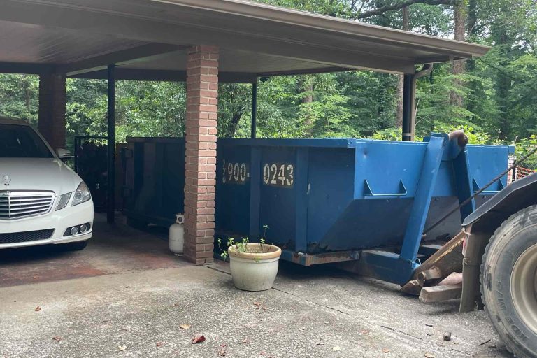 basement-cleanout-dumpster-rental-brookhaven-ga White Mercedes under a carport next to a large blue dumpster, with brick columns and greenery in the background.