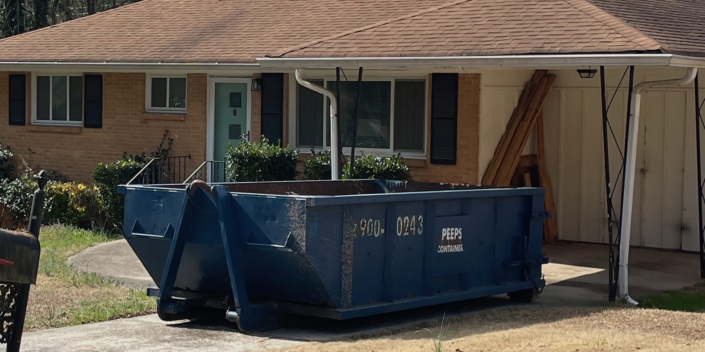 East point roof job 15 yard One-story brick house with a large blue dumpster in the driveway and wooden boards stacked beside a carport.