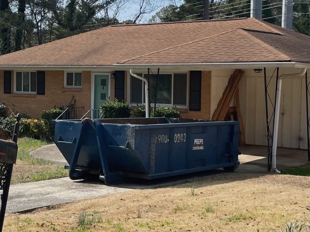 East point roof job 15 yard One-story brick house with a large blue dumpster in the driveway and wooden boards stacked beside a carport.
