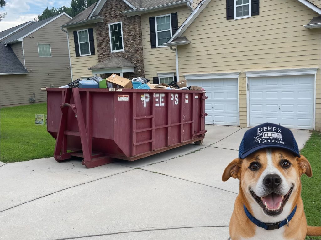 20 yard house clean up Fairburn - Peeps Containers Dog wearing a blue cap stands in a driveway next to a full red dumpster in front of a beige house with two white garage doors.