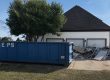 Blue dumpster parked in a driveway in front of a white house, with a pile of debris and metal scaffolding nearby under a clear blue sky.