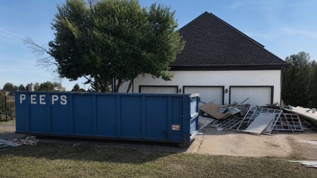 renovation-debris-dumpster-rental-fayetteville-ga Blue dumpster parked in a driveway in front of a white house, with a pile of debris and metal scaffolding nearby under a clear blue sky.