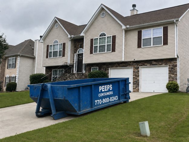 Residential Cleanout Dumpster Rental in Atlanta Blue dumpster labeled 'Peeps Containers' sits in the driveway of a beige two-story house with stone veneer and white garage doors.