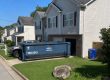 Blue garage cleanout dumpster in the driveway of a suburban home, with a grey SUV nearby and a blue recycling bin nearby on the lawn.