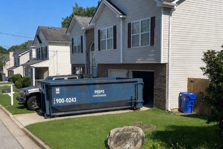 residential-garage-cleanout-dumpster-rental-atlanta-area Blue garage cleanout dumpster in the driveway of a suburban home, with a grey SUV nearby and a blue recycling bin nearby on the lawn.
