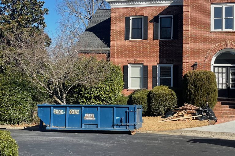 15 yard Smyrna flooring replacement Already had a picture from here Blue construction dumpster in a parking lot, with a brick multi-story building and piles of wood debris nearby.