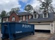 Blue dumpster on the driveway in front of a two-story brick-and-siding house with dormer windows. Placed near an open garage door.