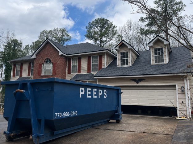 What Size Dumpster Do I Need? Blue dumpster on the driveway in front of a two-story brick-and-siding house with dormer windows. Placed near an open garage door.