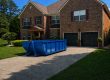 Blue dumpster parked in the driveway in front of a two-story brick house with a two-car garage on a sunny day.