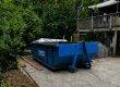 Blue dumpster labeled 'Peeps Containers' on a concrete driveway with wooden planks nearby and a wooden deck in a green, partially wooded yard.
