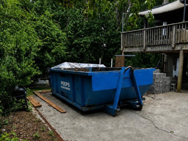 sandy-springs-moving-cleanout-dumpster-rental Blue dumpster labeled 'Peeps Containers' on a concrete driveway with wooden planks nearby and a wooden deck in a green, partially wooded yard.