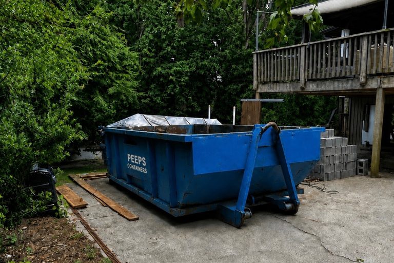 sandy-springs-moving-cleanout-dumpster-rental Blue dumpster labeled "PEEPS Containers" sits on a concrete pad beside a wooden deck, with lumber and concrete blocks nearby in a backyard renovation area.